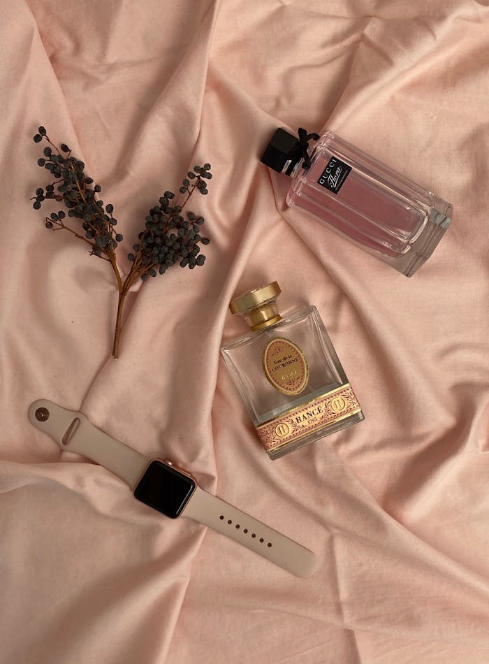 A stylish flat lay of perfume bottles, a smartwatch, and dried leaves on soft pink fabric.